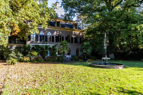 Historisch landhuis aan de Langbroekerdijk met gotische ramen, omringd door herfstige bomen en een sierlijke fontein in de tuin.